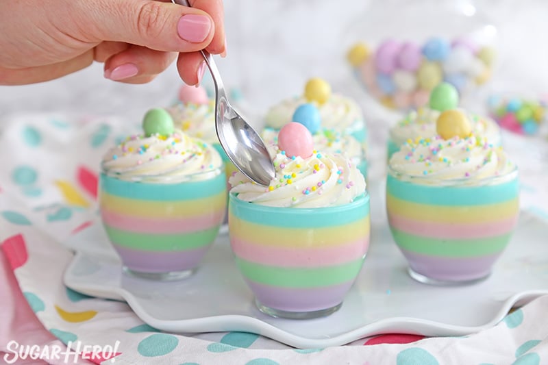 Seven pastel rainbow gelatin cups, one with a spoon hovering above it.