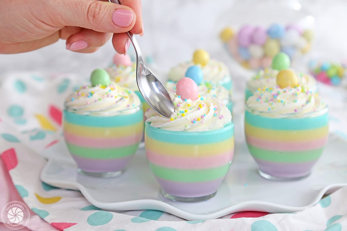 Seven pastel rainbow gelatin cups, one with a spoon hovering above it.