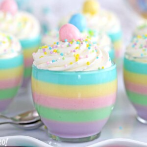 Seven Pastel Rainbow Gelatin Cups on a white plate with scalloped edges and two metal spoons to the side of the cups.