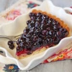 A slice of blueberry coconut tart in a square white dish and a fork set beside it.