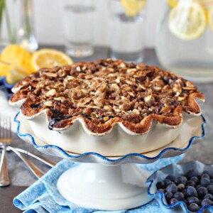 Blueberry crumble pie on a white cake stand with scalloped edges and fresh blueberries and lemons in the background.
