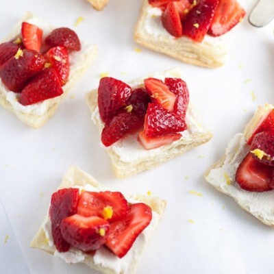 Five honeyed strawberry and mascarpone tartlets on a white background with lemon zest sprinkled around.