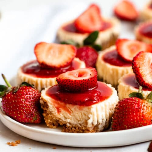 Six mini strawberry cheesecakes with fresh strawberries scattered on a round white plate.