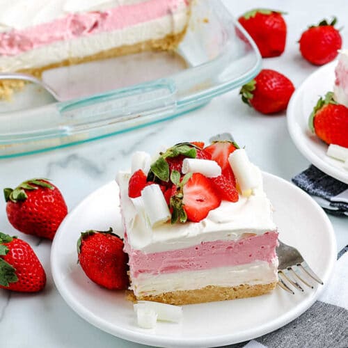 A slice of strawberry lasagna on a small, round, white plate with fresh strawberries in the background.