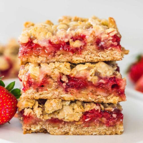 Three strawberry oatmeal bars stacked on top of each other with a few fresh strawberries in the background.
