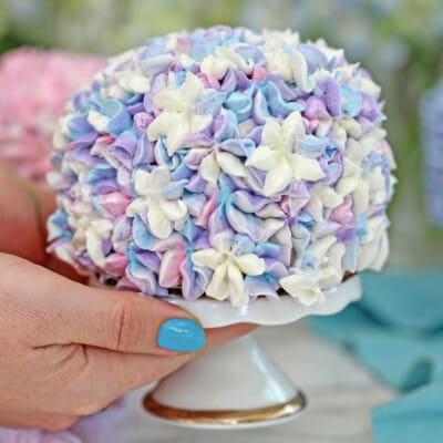 Hand holding a mini cake stand with a hydrangea cake on it.