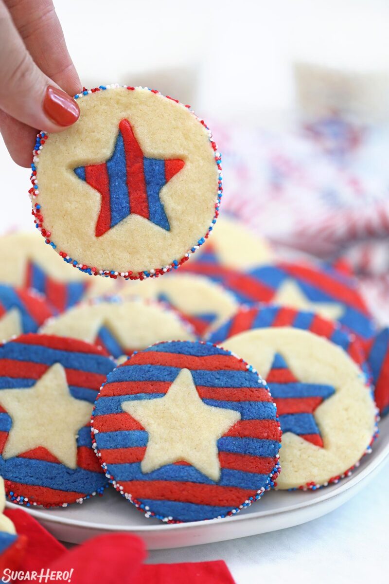 Hand holding up a Stars and Stripes Sugar Cookie from a plate of other cookies.