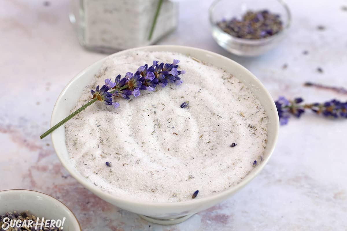 Bowl of lavender sugar on marble background, with fresh lavender scattered on top and around.