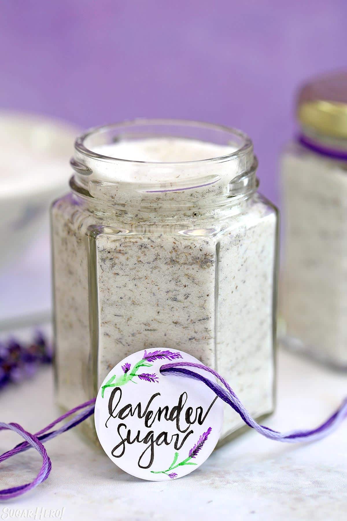 Hexagonal glass jar filled with lavender sugar, on a marble surface with a purple background.