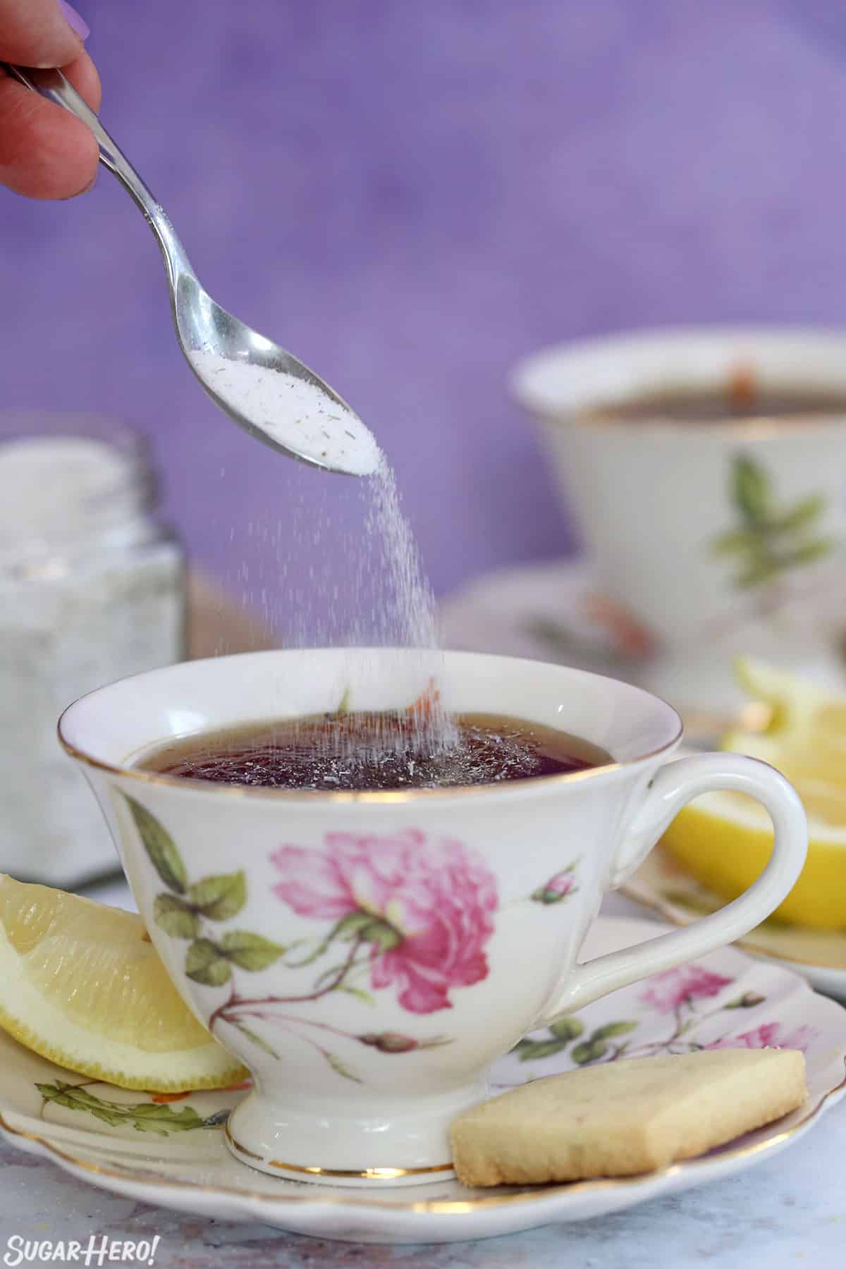 Spoon sprinkling lavender sugar into a floral teacup, with a lemon wedge and shortbread cookie on the side.