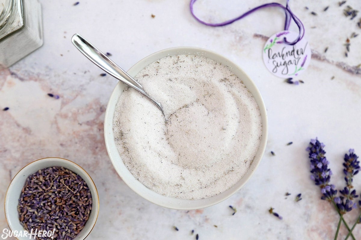 Overhead shot of white bowl filled with lavender sugar on a marble surface, with fresh lavender scattered around.