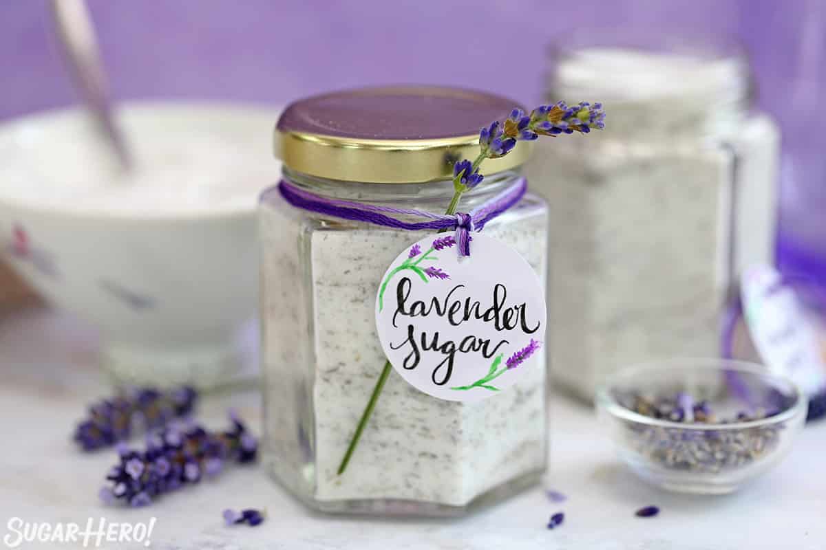 Hexagonal jar of lavender sugar with a gold lid, handwritten label, and fresh sprig of lavender tied around.