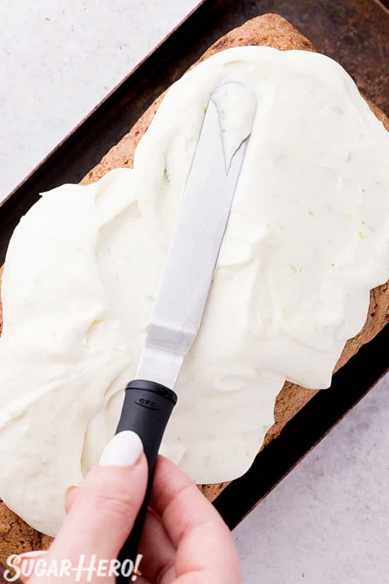 Hand holding a mini spatula, spreading cream cheese frosting on a loaf of zucchini bread.