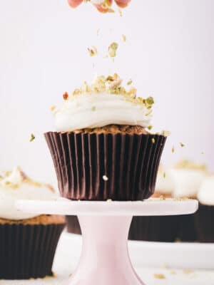 Hand sprinkling crushed pistachios on top of a zucchini cupcake frosted with cream cheese frosting.