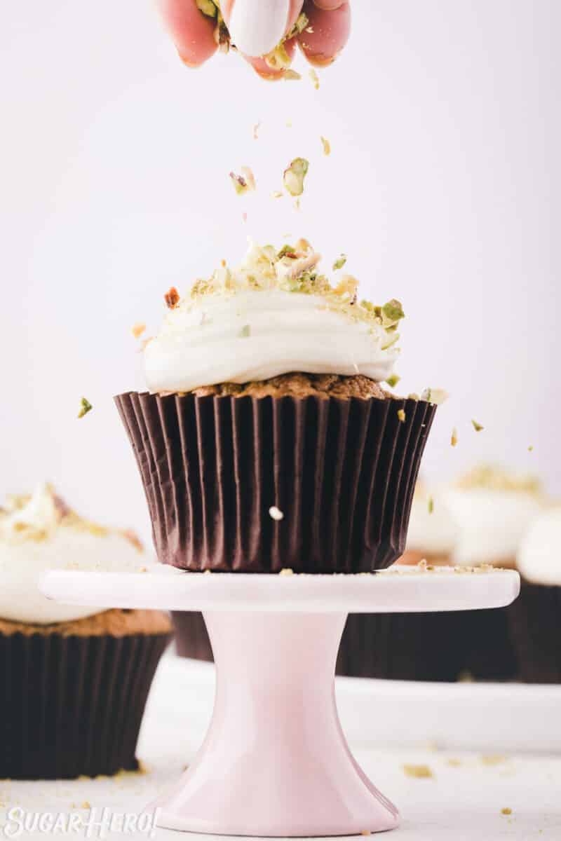 Hand sprinkling crushed pistachios on top of a zucchini cupcake frosted with cream cheese frosting.