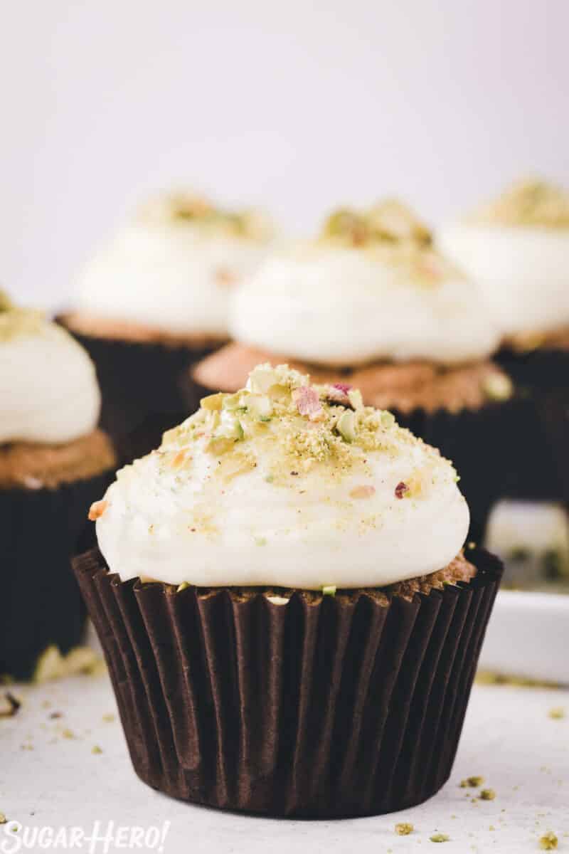Zucchini cupcake in a dark brown wrapper, with cream cheese frosting and crushed pistachios on top.