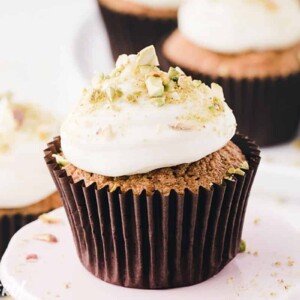 Zucchini Cupcake in brown liner on a white cupcake stand, topped with cream cheese frosting and crushed pistachios.