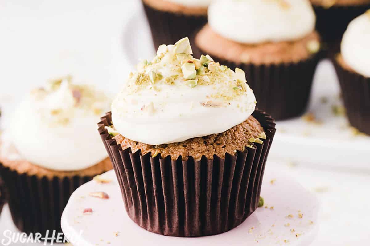 Zucchini Cupcake in brown liner on a white cupcake stand, topped with cream cheese frosting and crushed pistachios.