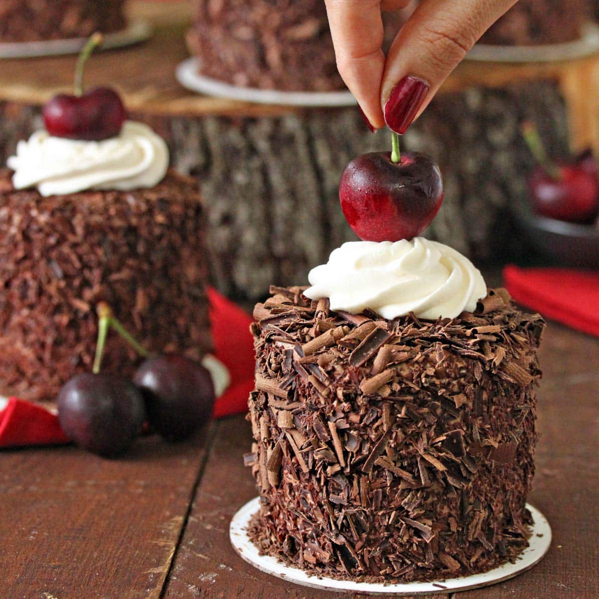 Close up of black forest mini cake with hand placing cherry on top and additional mini cakes in the background.