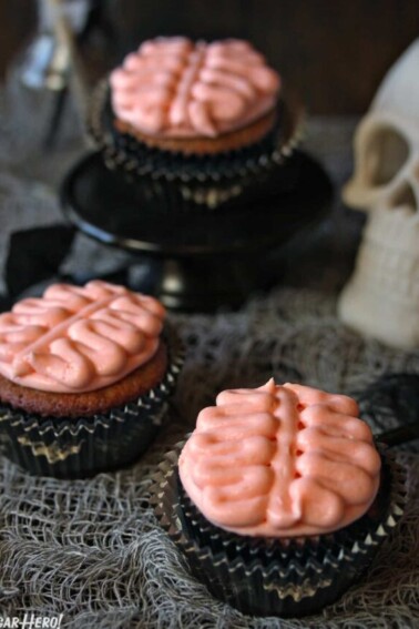 3 Brain Cupcakes next to a skull on a grey webbed tablecloth.