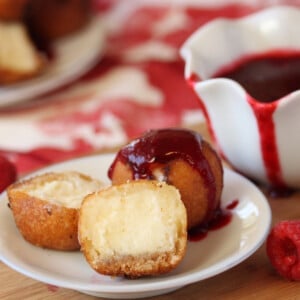 1 deep-fried cheesecake bite cut to show center and another drizzled with raspberry sauce on a white plate with sauce in the background.