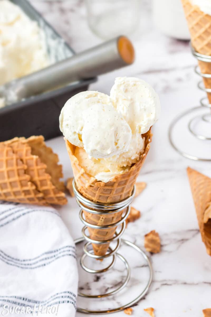 Waffle cone with scoops of vanilla ice cream, with more ice cream and waffle cones in the background.