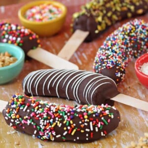 Assortment of chocolate-dipped frozen bananas with sprinkles and white chocolate on a wooden surface.