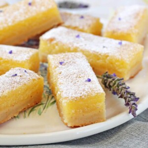 Seven lavender lemon bars on a round white plate with a lavender flower next to them.