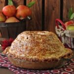 Mile High Apple Pie with crisp golden crust on a red napkin with baskets of apples in the background.