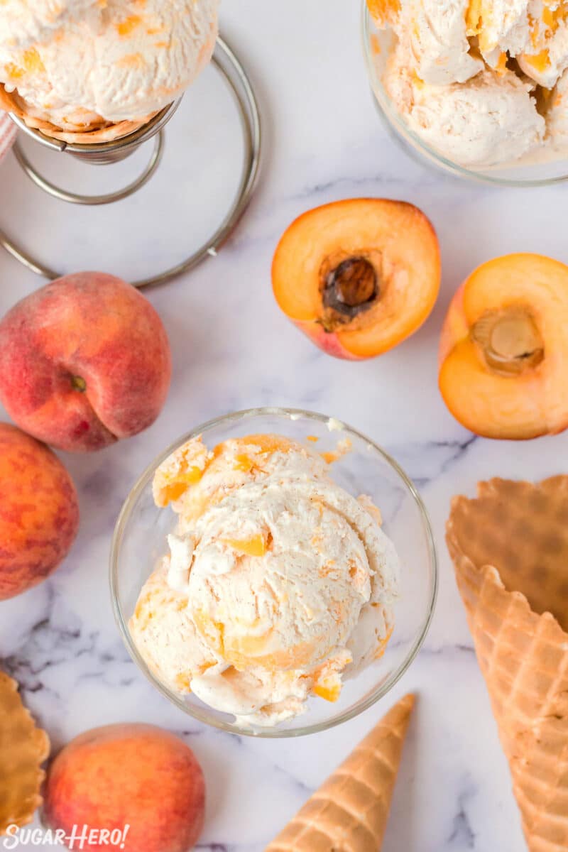 Top view of No-Churn Peach Ice Cream in a clear glass bowl with peaches and waffle cones in the background.