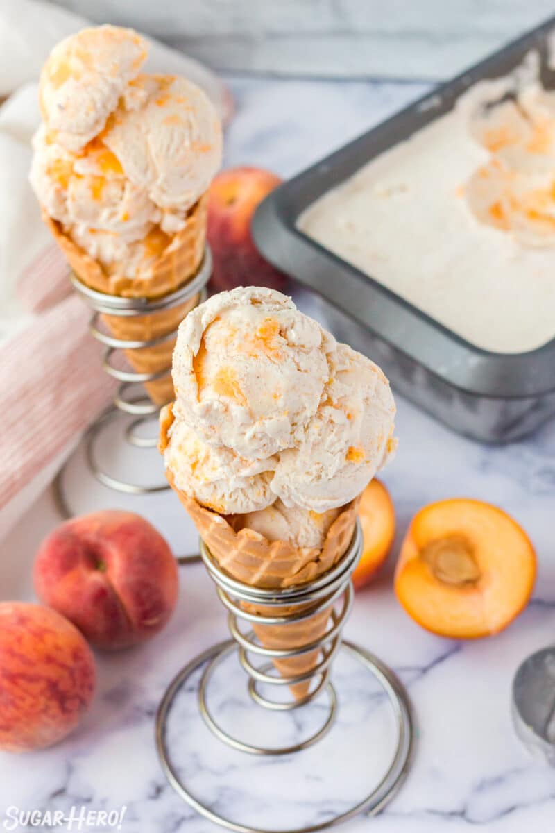 Top view of 2 waffle cones filled with scoops of No-Churn Peach Ice Cream in silver cone holders with peaches in the background.