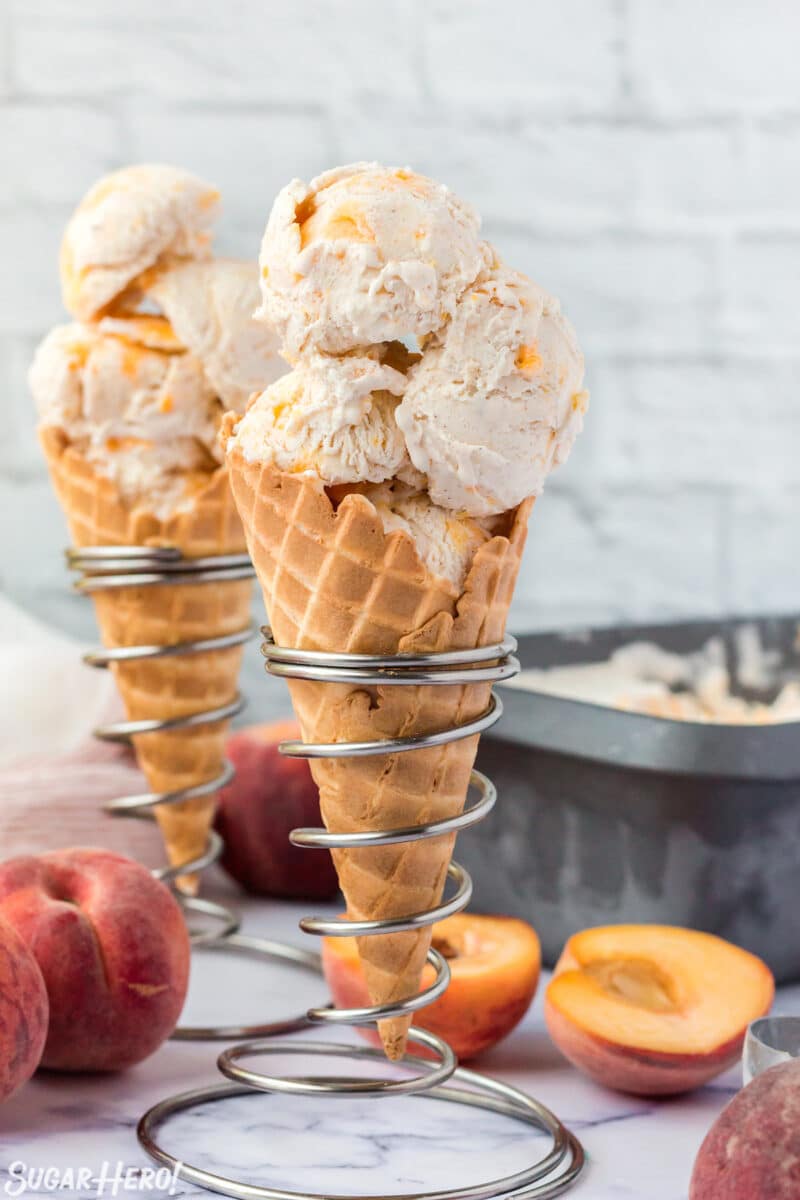 2 waffle cones filled with scoops of No-Churn Peach Ice Cream in silver cone holders with peaches in the background.