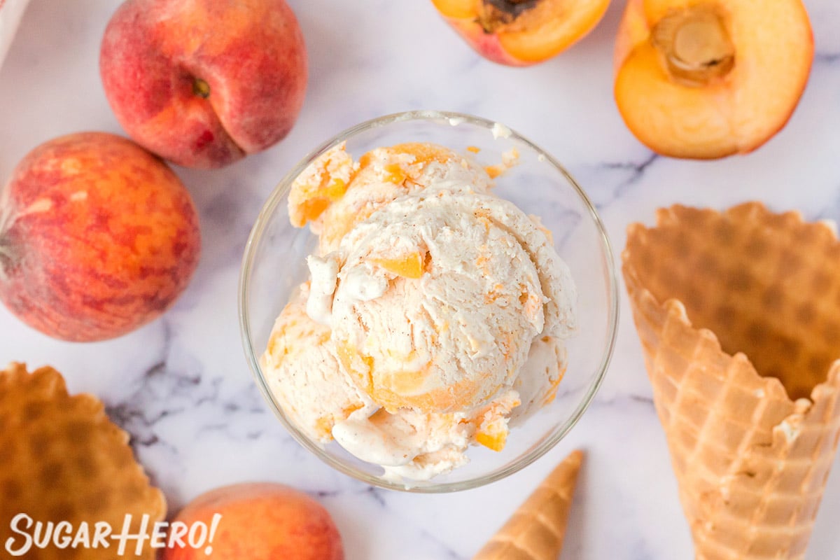Top view of No-Churn Peach Ice Cream in a clear glass bowl with peaches and waffle cones in the background.