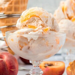 3 Scoops of No-Churn Peach Ice Cream in a clear glass bowl with peaches and waffle cones in the background.