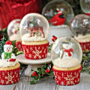 Several snow globe cupcakes displayed with holly on a wooden surface.