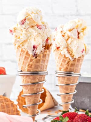 2 waffle cones filled with scooped No-Churn Strawberry Shortcake Ice Cream with strawberries in the background.