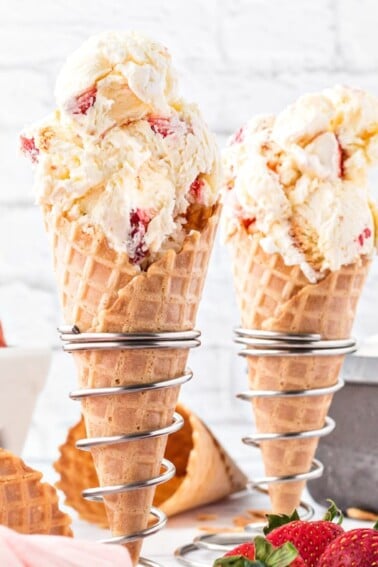 2 waffle cones filled with scooped No-Churn Strawberry Shortcake Ice Cream with strawberries in the background.