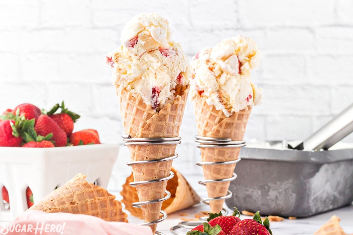 2 waffle cones filled with scooped No-Churn Strawberry Shortcake Ice Cream with strawberries in the background.