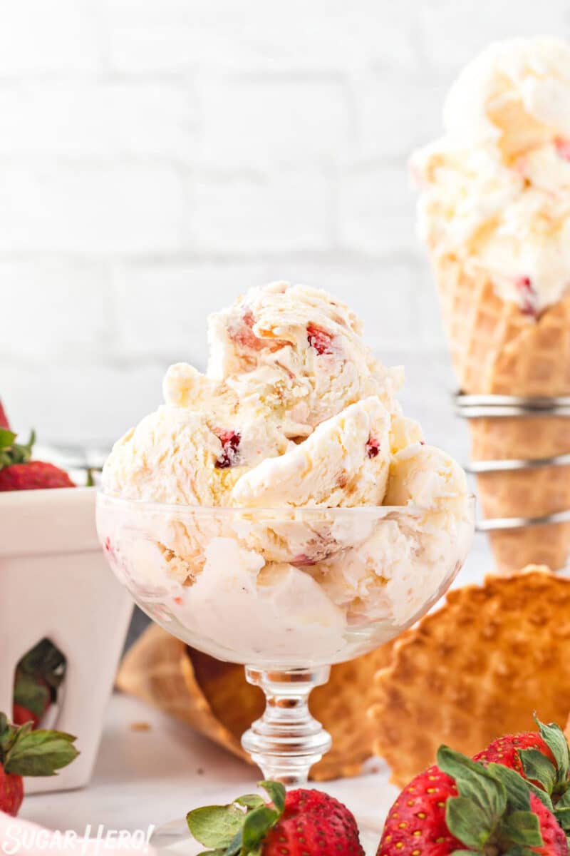 3 Scoops of No-Churn Strawberry Shortcake Ice Cream in a clear glass bowl with strawberries and waffle cones in the background.