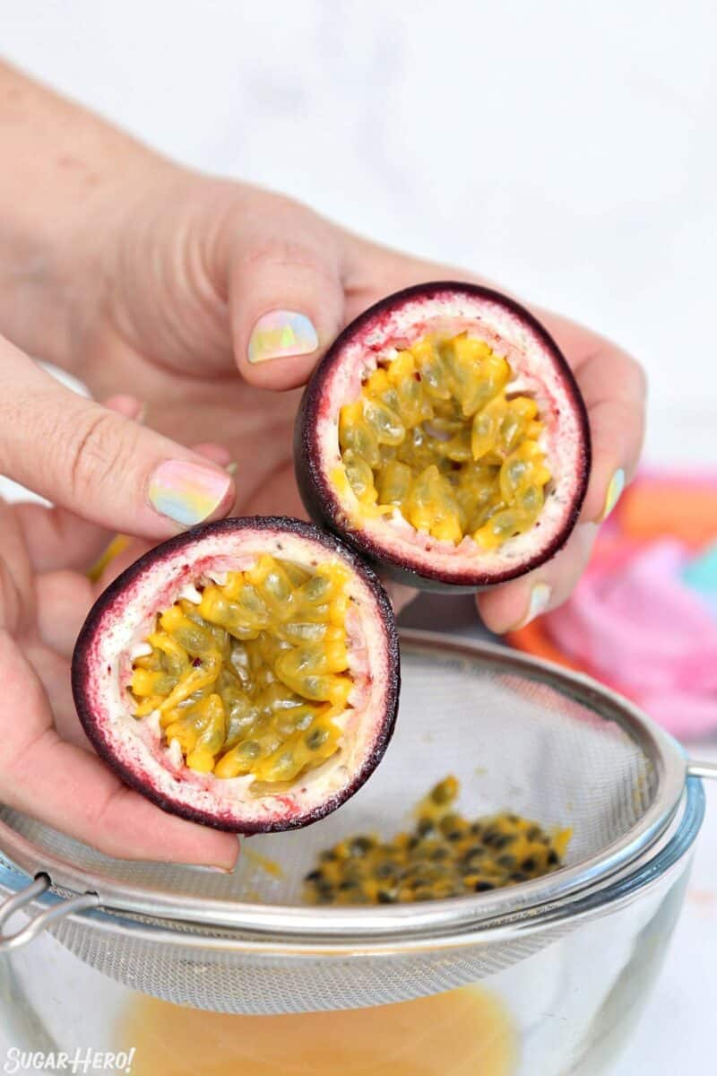 Hands holding two cut passion fruit halves over a wire mesh strainer.