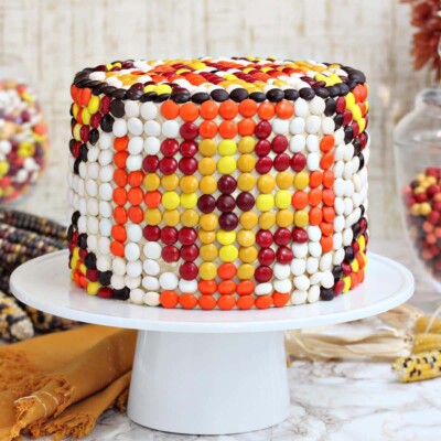 Patterned M&M Cake on a white cake plate next to an orange napkin and dried corn.