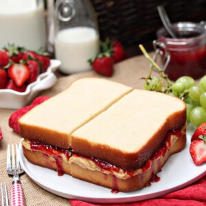 Peanut Butter and Jelly Sandwich Cake on a white plate with fruit, jam and milk in the background.