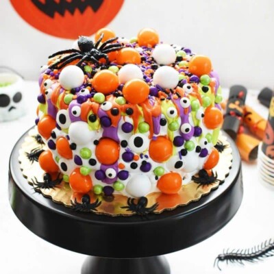 Layer cake decorated with orange, green, and purple frosting drips, candy, and candy eyeballs.