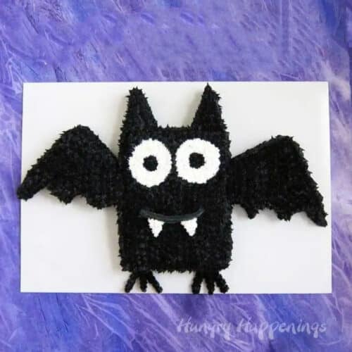 A black sheet cake in the shape of a bat with black and white piping, on a white background bordered by purple.