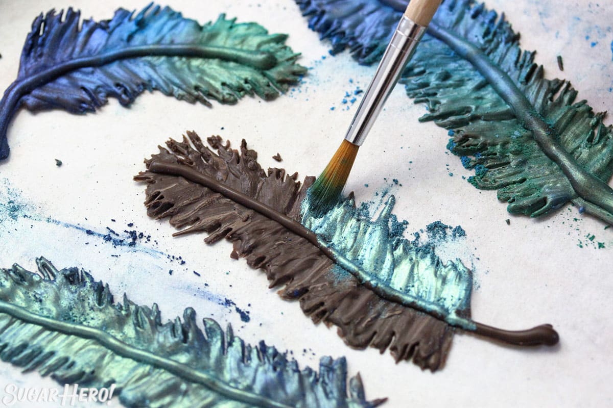Brushing teal luster dust on a chocolate feather, with blue and green chocolate feathers scattered around.
