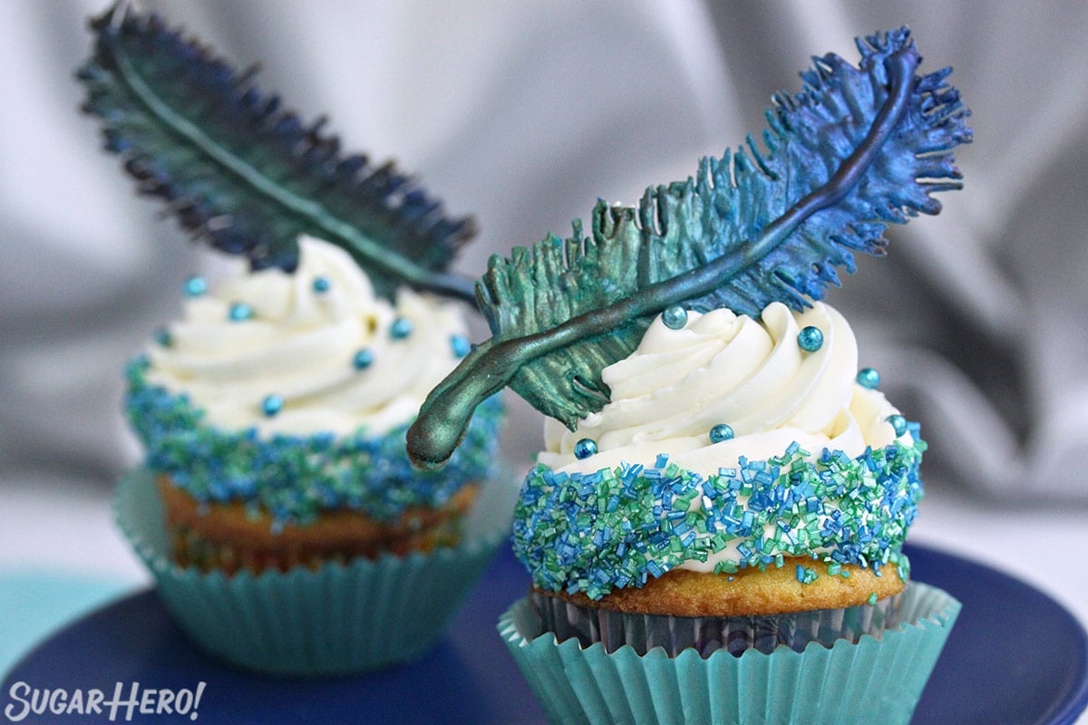 Two cupcakes on a blue plate with blue-green chocolate feathers on top.