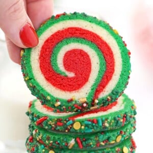 Hand with red fingernail lifting up a Christmas Pinwheel Cookie from a stack of cookies.