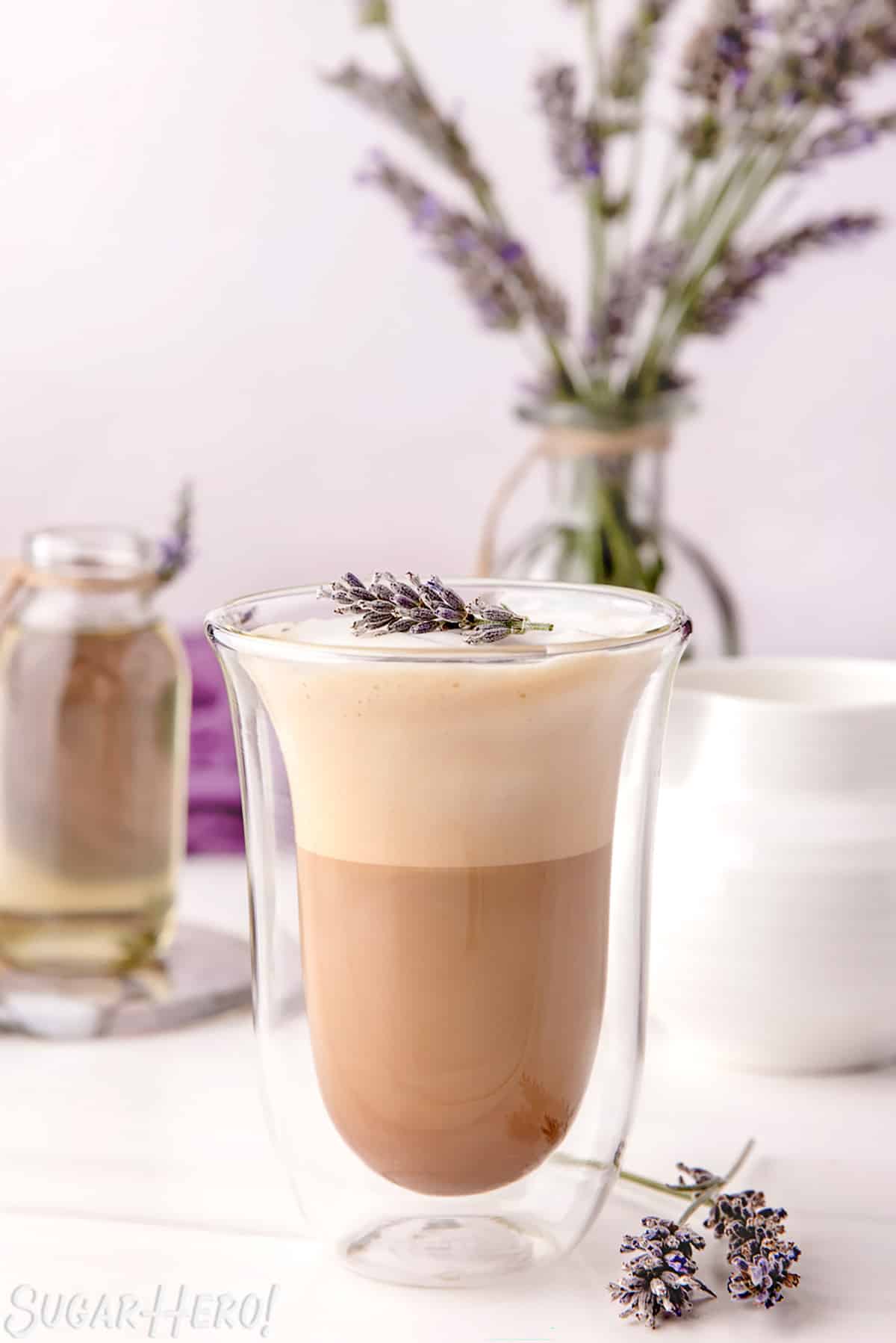 Lavender Latte in a clear glass jar, with simple syrup and a vase of fresh lavender in the background.