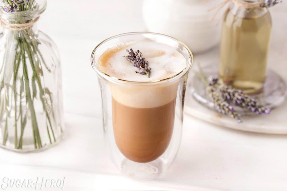 Lavender Latte in a glass, with simple syrup and a vase of fresh lavender in the background.