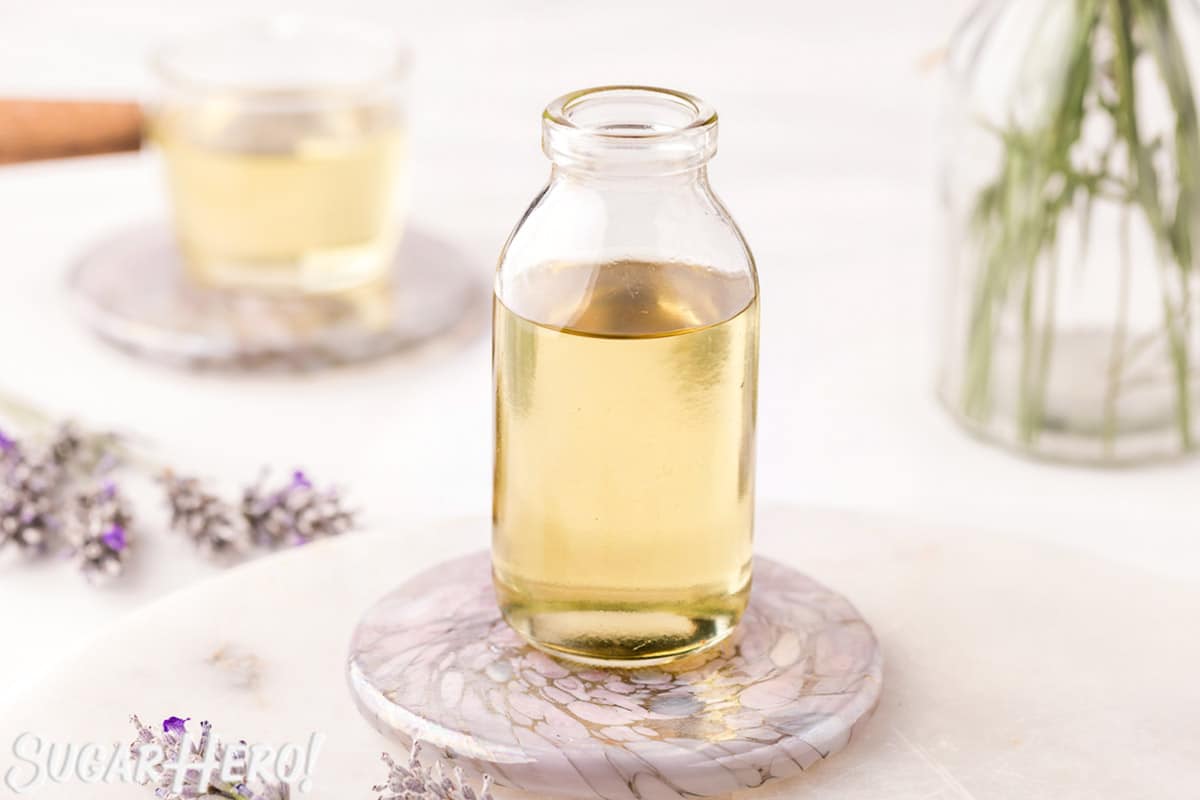 Lavender syrup in a clear glass jar, with fresh lavender and more simple syrup in the background.